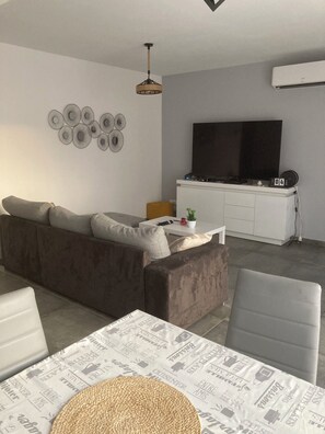A modern living room with a sofa, a dining table with a placemat, a hanging light fixture, and a flat-screen TV on a white cabinet.
