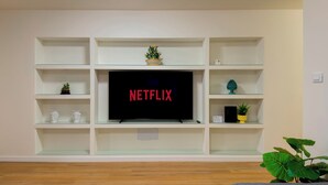 Een woonkamer met een televisie aan een witte muur, omringd door glazen schappen met decoratieve objecten.