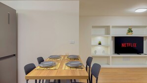 Een moderne woonkamer met een eettafel voor vier personen, een flatscreen-tv waarop 'NETFLIX' wordt weergegeven, en een in de muur geïntegreerde kast met decoratieve objecten.