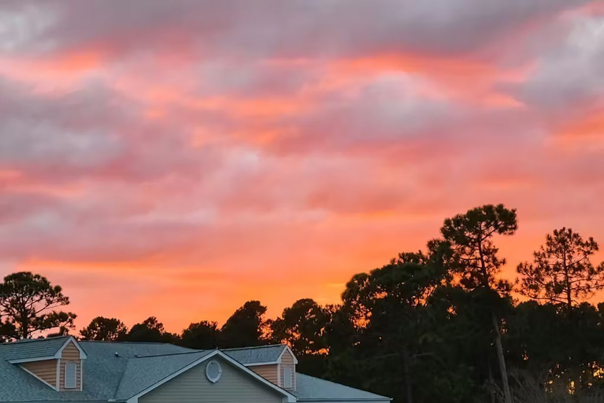 Una casa con tetto a spiovente e camino, circondata da alberi e cielo al tramonto.