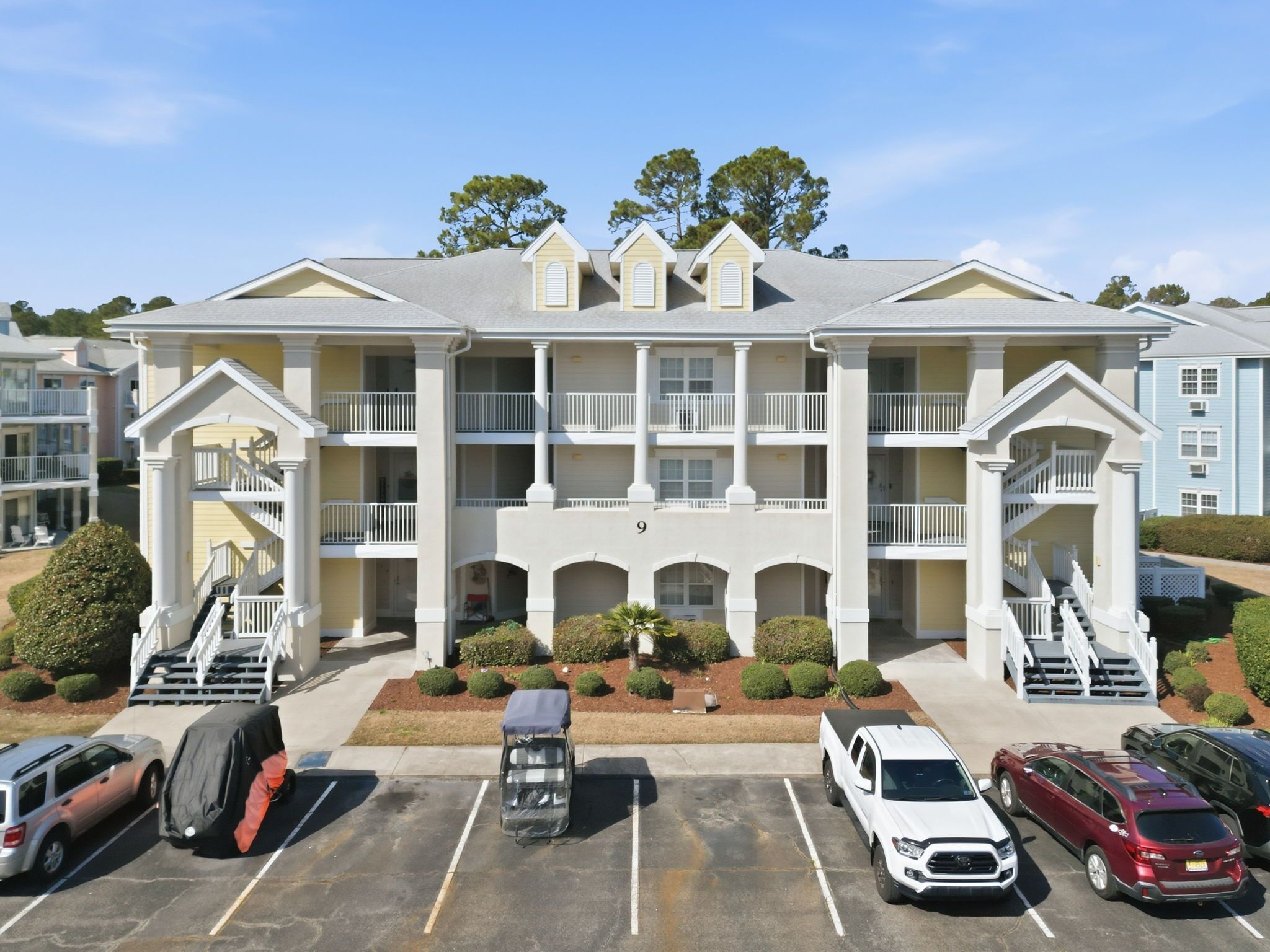 A multi-story hotel with a central courtyard, balconies on each floor, and a parking lot with several cars.