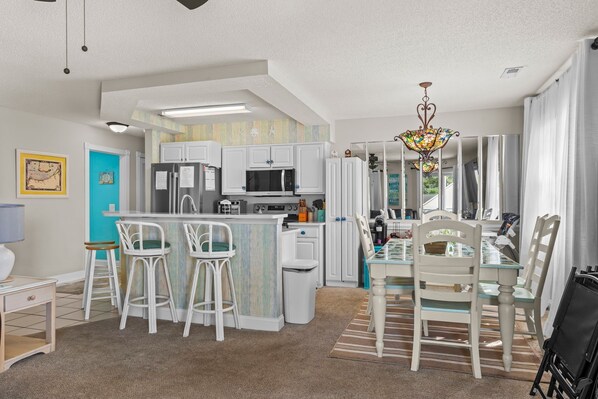 A modern kitchen with white cabinets, a central island with stools, and a dining area with a glass table and chairs. - Calabash Golf Condo | 15 Min to Beach (Calabash)