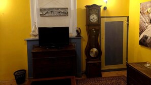 A living room with a television on a wooden stand, a large wall clock, and a painting on the wall. - Château Montegut - Private apartment in a medieval tower with Pyrenees view (Montégut)
