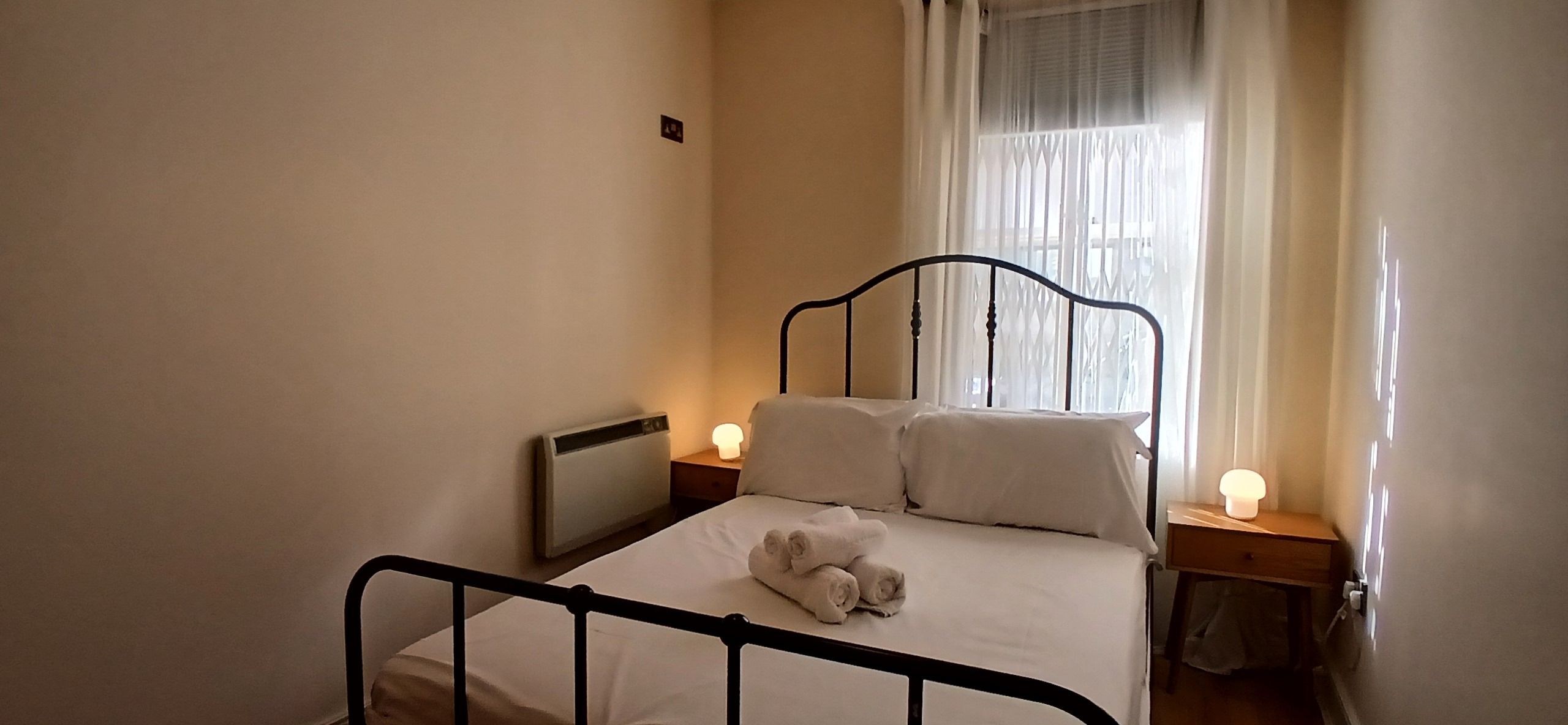 A single bed with a metal frame, two bedside tables with lamps, a radiator, and a window with sheer curtains.