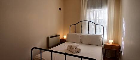 A single bed with a metal frame, two bedside tables with lamps, a radiator, and a window with sheer curtains.