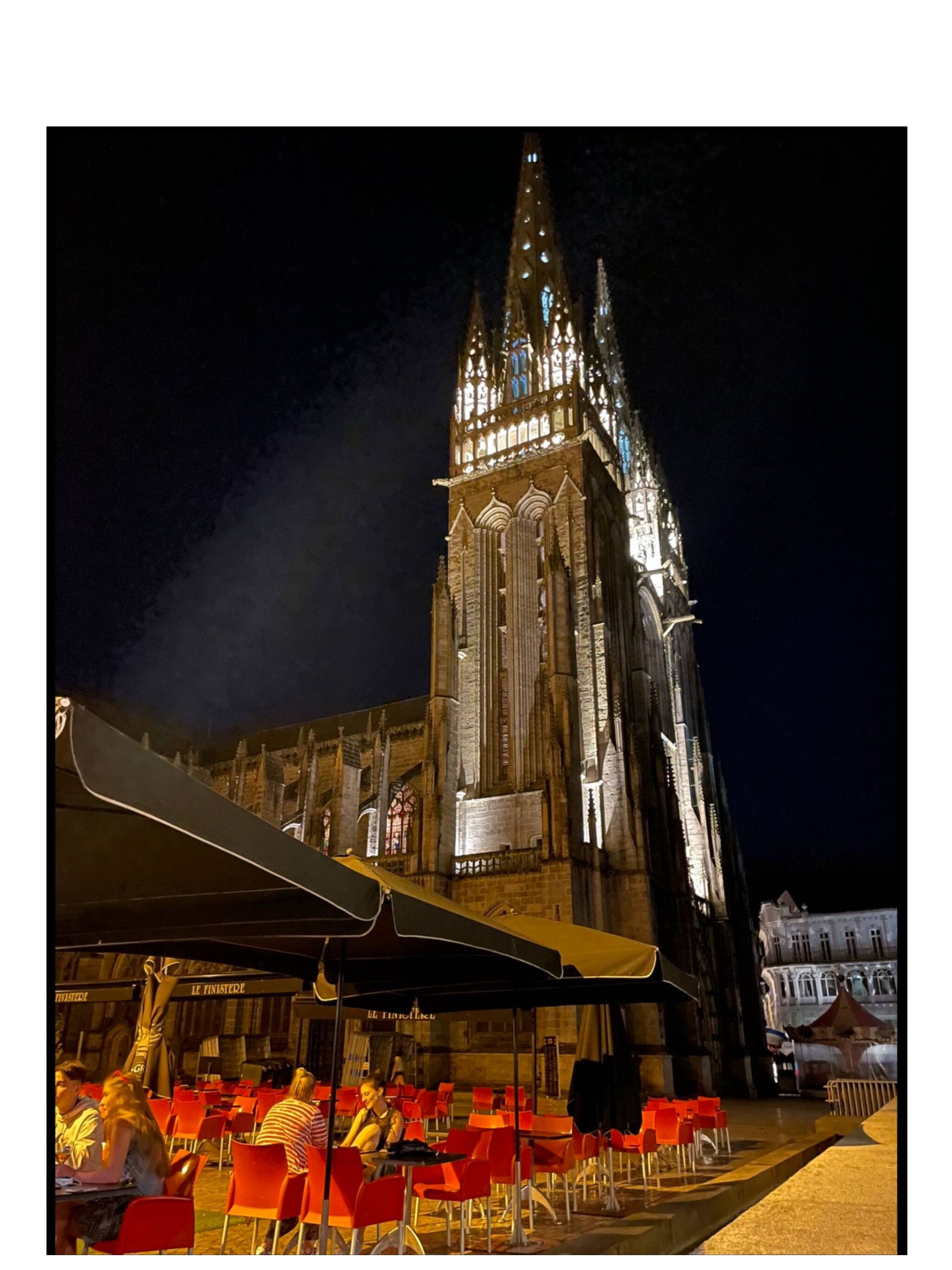 A historic building with a tall spire, a covered outdoor seating area with red chairs, and a sign reading "LE FINISTE."