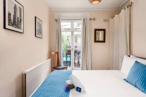 Een hotelkamer met een bed, een radiator, een stoel, een raam met gordijnen en afbeeldingen in lijsten aan de muur.