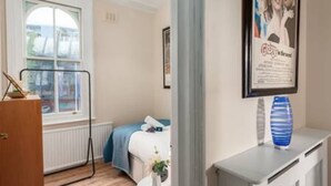 Une chambre d’hôtel avec un lit, une fenêtre, un radiateur, une petite table avec un vase et un tableau encadré au mur.