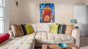 Un salon avec un canapé beige, des coussins colorés, un tableau au mur et une lampe sur une table d’appoint.