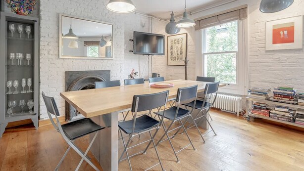 A dining area with a wooden table, black chairs, and pendant lights. A television is mounted on the wall, and there is a bookshelf with books and a framed picture. - Experience London in Amazing Reconverted Gin House (London)