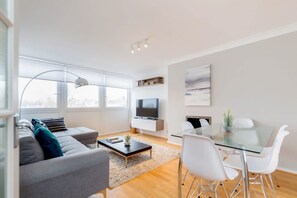 Un salon moderne avec un canapé gris, une table à manger en verre, des chaises blanches, un téléviseur à écran plat et une grande fenêtre.