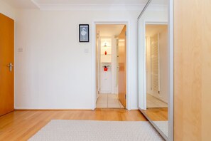 A hallway with a wooden floor, a white rug, and a door leading to a room with a red object on a shelf. - Bright 2bed Flat in Holland Park-notting Hill (London)