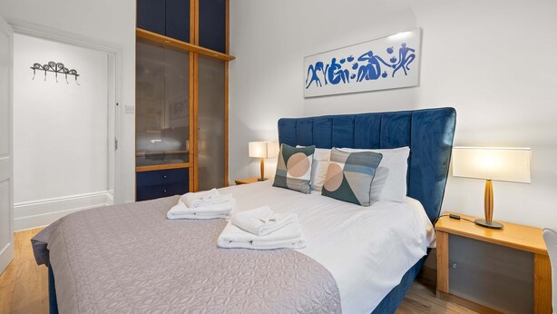 A neatly made bed with folded towels, a bedside lamp, and a wall art piece. - Beautiful 2bed Flat With Balcony in Earls Court (London)