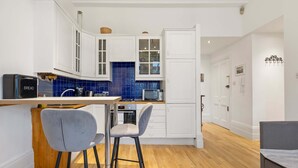 A modern kitchen with white cabinets, a blue-tiled backsplash, and a wooden countertop. There are two grey bar stools and a small dining table. - Beautiful 2bed Flat With Balcony in Earls Court (London)