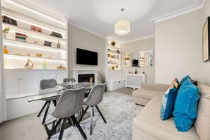 A modern living room with a glass dining table, grey chairs, a beige sofa, and a fireplace. - Great Central Location - 2bed Chelsea Earls Court (London)