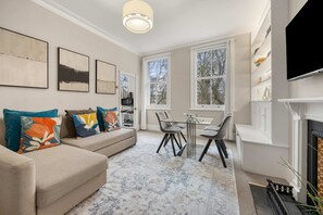 A living room with a sofa, a dining table, and chairs. There are large windows and a fireplace. - Great Central Location - 2bed Chelsea Earls Court (London)