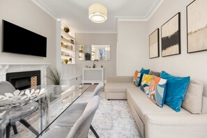 A modern living room with a flat-screen TV, a glass-top dining table, a beige sofa with colorful pillows, and a fireplace. - Great Central Location - 2bed Chelsea Earls Court (London)