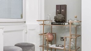 A modern living room with a round marble table, grey ottomans, a bookshelf with decorative items, and a framed artwork on the wall. - Beautiful Refurbished 2bed 2bath Next to Hyde Park (London)