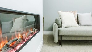 A modern living room with a glass fireplace, a grey sofa with white cushions, and a carpeted floor. - Beautiful Refurbished 2bed 2bath Next to Hyde Park (London)