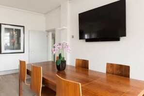 A modern dining area with a wooden table and chairs, a vase with flowers, and a mounted flat-screen TV on the wall. - Modern Stylish 2bed 2bath Close to Kings Road (London)