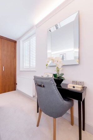 A bedroom with a wooden wardrobe, a bedside table with a mirror, a grey upholstered chair, and a potted orchid. - Modern Stylish 2bed 2bath Close to Kings Road (London)