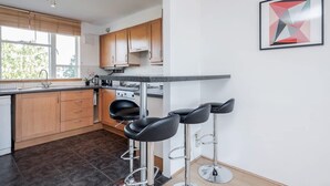 A modern kitchen with wooden cabinets, a built-in oven, and a large window. There are three black bar stools and a framed abstract painting on the wall. - Spacious 2bed in top Location Holland Park-n Hill (London)