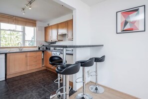 A modern kitchen with wooden cabinets, a built-in oven, and a large window. There are three black bar stools and a framed abstract painting on the wall. - Spacious 2bed in top Location Holland Park-n Hill (London)