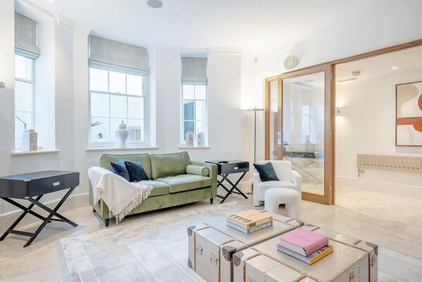 A modern living room with a green sofa, white armchair, and a coffee table with books. There are large windows and a sliding glass door leading to a balcony. - Spacious Modern 2bed 2 5bath in Pimlico-victoria (London)