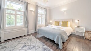 A bedroom with a bed, bedside tables, a chandelier, and large windows with curtains. - Stylish 3-floor 3bed Home With Garden Near River (London)