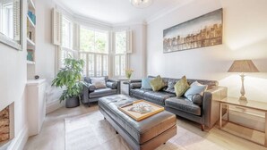 A modern living room with a fireplace, leather sofas, a coffee table, and a cityscape painting. - Stylish 3-floor 3bed Home With Garden Near River (London)