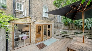 A courtyard with a wooden deck, a dining area with glass doors, and a large umbrella. - Stylish 3-floor 3bed Home With Garden Near River (London)
