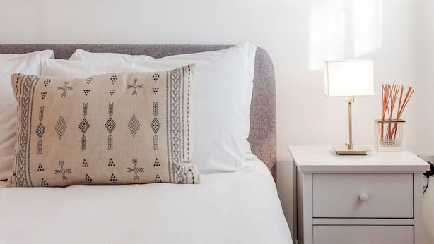 Un lit bien fait, agrémenté d’un coussin à motifs, d’une lampe de chevet et d’un petit récipient contenant des bâtonnets de rotin.