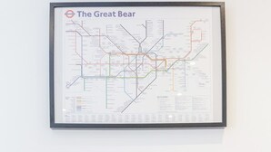 Eine gerahmte U-Bahnkarte mit dem Titel „Der Große Bär“ an einer Wand, ein Holztisch mit Schublade und drei gerahmte Zertifikate darunter.