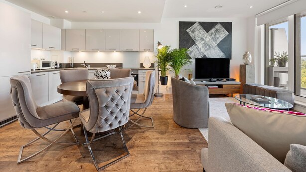 A modern kitchen with white cabinets, a dining area with grey upholstered chairs, and a living room with a grey sofa and a TV. - Luxury and Quiet Oasis in London (London)