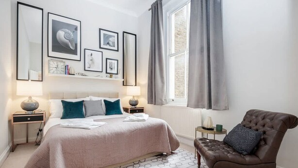 A bedroom with a bed, two bedside tables, a chair, a window with curtains, and wall-mounted artwork. - Charming Spacious 1bed Near Portobello Rd N Hill (London)