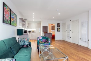 A modern living room with a green sofa, a glass coffee table, and a colorful armchair. There is a kitchen area in the background with white cabinets and a dining table. - Modern and Large Oasis Flat in Camden With Patio (London)