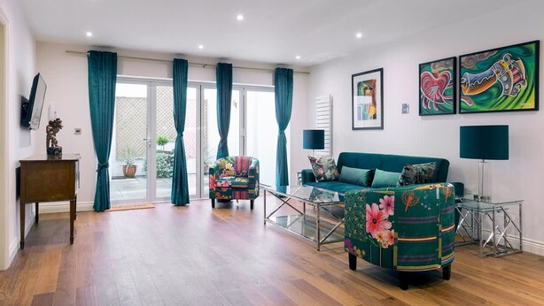 Un salón amplio con sofá, sillón y mesa de centro, una puerta corrediza de vidrio que da a un balcón, y obras de arte en las paredes.