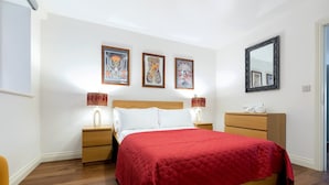 Un dormitorio con una cama de madera, una colcha roja, dos lámparas de noche, una mesita de noche, un aparador y tres cuadros en la pared.