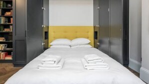 A neatly made bed with white linens and pillows, a yellow headboard, and a bookshelf to the left. - Modern Spacious Studio in Chelsea by King s Road (London)