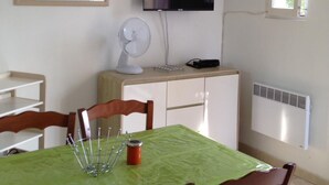 Ein Zimmer mit Esstisch, der mit einer grünen Tischdecke bedeckt ist, Holzstühlen, einem weißen Deckenventilator, einem an der Wand montierten Flachbildfernseher, einem Fenster mit Blick ins Grüne und einem Heizkörper an der Wand.