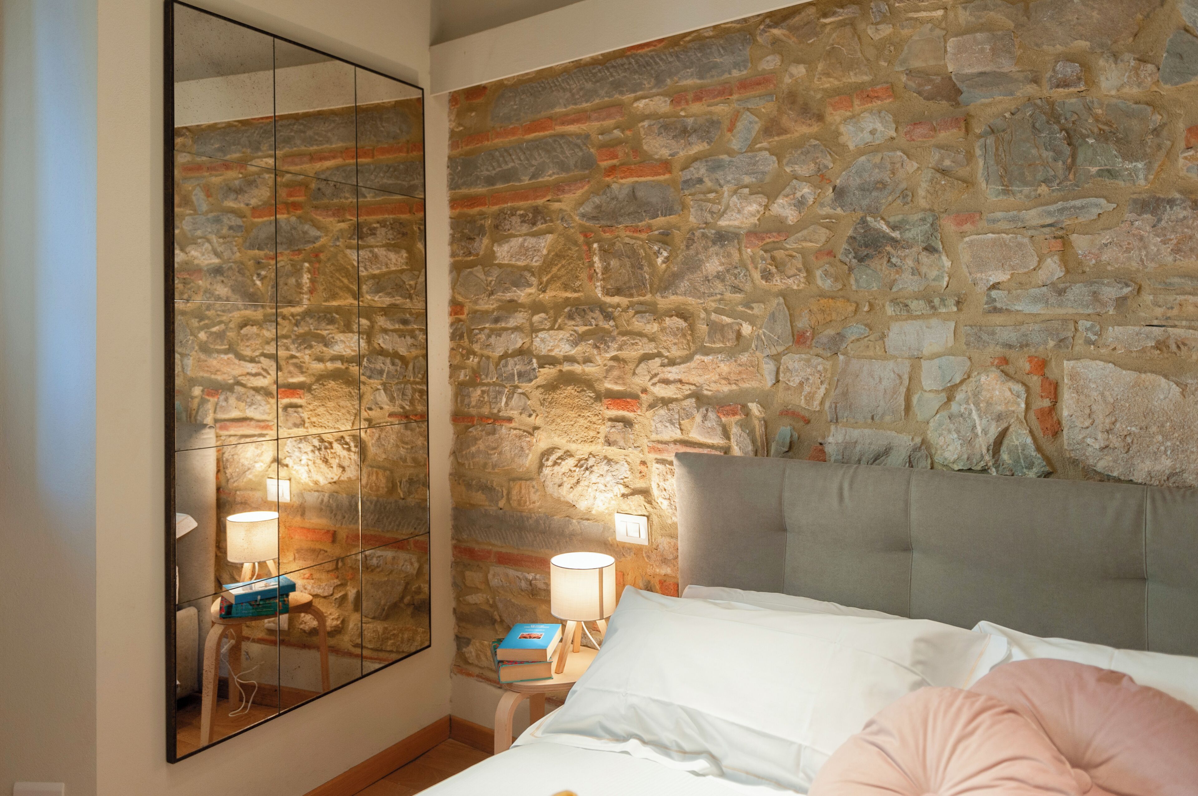 Un dormitorio con pared de piedra, un espejo grande, una cama con almohadas y lámparas de noche.