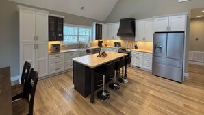 Uma cozinha espaçosa com armários brancos, uma ilha grande com banquetas, uma geladeira de aço inoxidável e uma janela que proporciona luz natural.