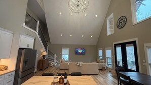 Uma sala de estar espaçosa com teto alto, um lustre, uma televisão grande e uma mesa de jantar com uma bandeja de garrafas.