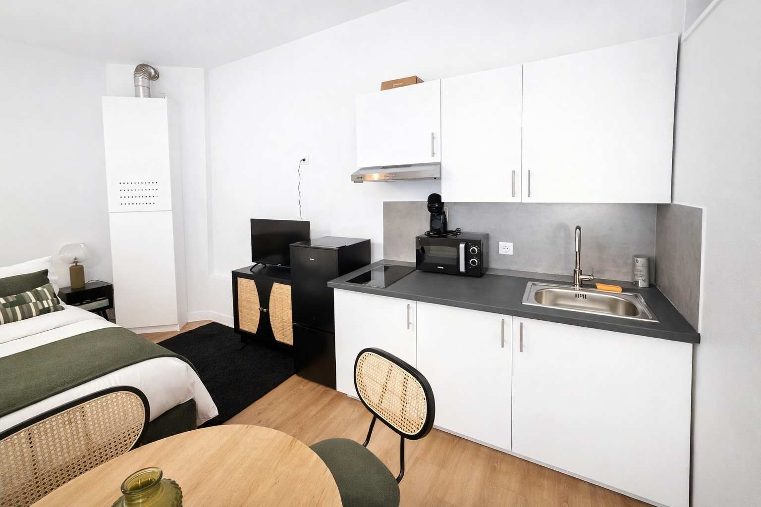 Habitación de hotel pequeña con kitchenette, una cama con ropa de cama verde, una pequeña mesa de comedor y una silla.