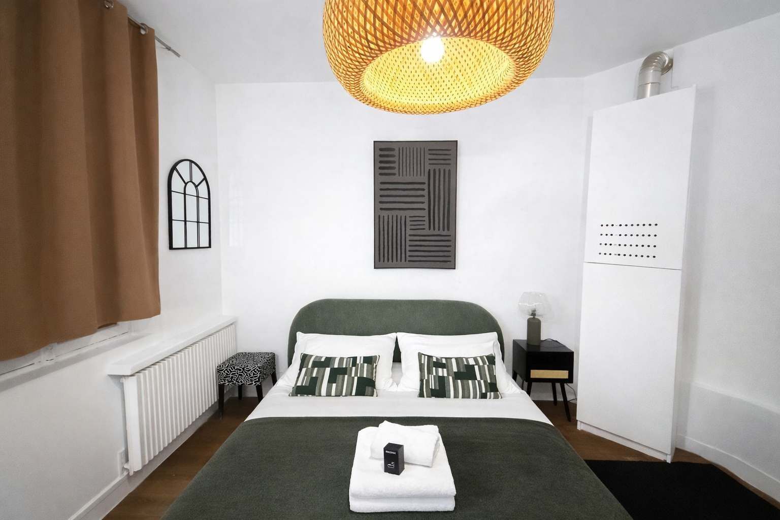 Une chambre bien rangée, avec un lit, un radiateur, une fenêtre avec des rideaux, une œuvre d’art accrochée au mur, une table de chevet avec une lampe et une armoire.