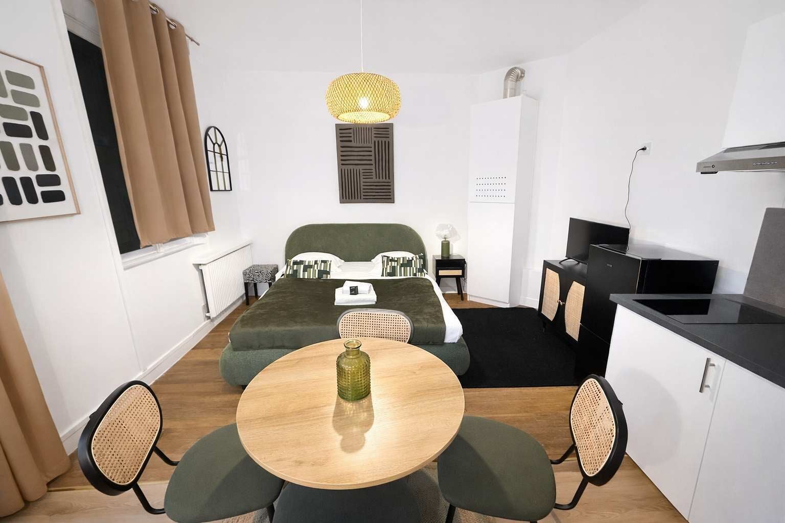 A compact hotel room with a bed, a dining table with chairs, a kitchenette, and a hanging light fixture.