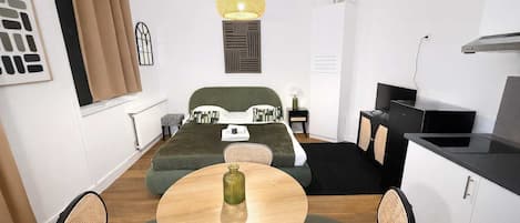 A compact hotel room with a bed, a dining table with chairs, a kitchenette, and a hanging light fixture.