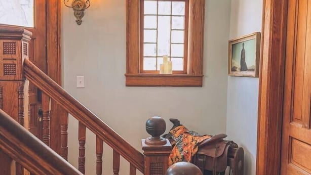 Un escalier en bois, doté d’une balustrade, mène à un palier. On aperçoit une fenêtre avec un cadre en bois et un luminaire fixé au mur.