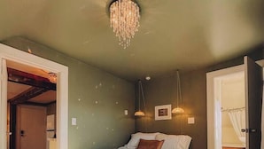 Une chambre avec un plancher en bois, un lit, un lustre et une porte donnant sur une autre pièce.