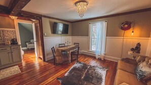 Un salon chaleureux avec un plancher en bois, un coin repas avec une table et des chaises, un téléviseur fixé au mur et un lustre.